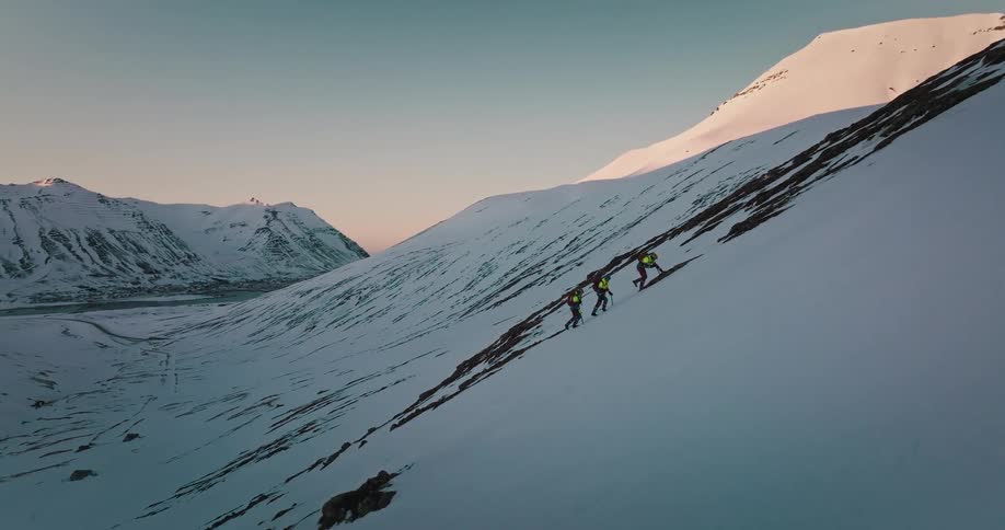 Aerial view of three mountaineers ascending a steep snowy mountain ridge with ice axes as the camera pulls back to reveal a vast valley at dawn
