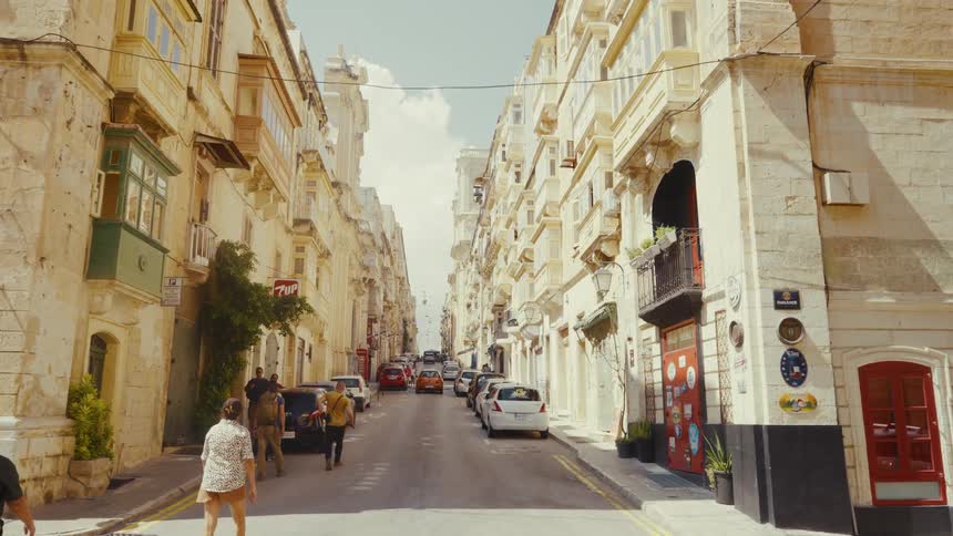 A slow motion point-of-view walk down a sunny street in Valletta, Malta, lined with historic sandstone buildings and parked cars