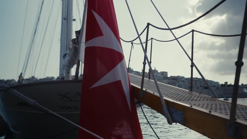 A red flag with a white Maltese cross hangs from a classic sailboat docked in the Valletta harbor in Malta