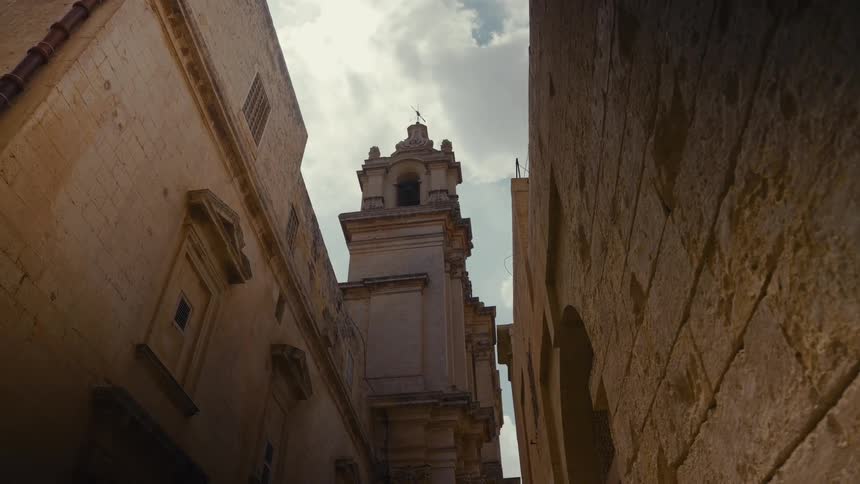 A low-angle view from a narrow stone alleyway looks up at an ancient church bell tower against a cloudy sky