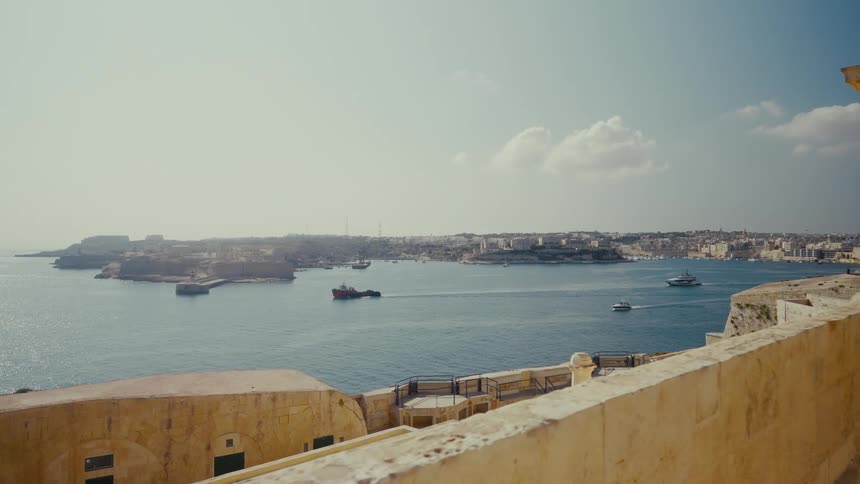 A scenic dolly shot from a fortress wall overlooks the bustling Grand Harbour of Valletta, Malta, with boats and ships navigating the blue waters on a sunny day