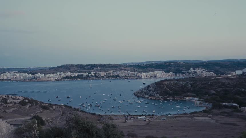 An airplane flies over a tranquil bay filled with boats as evening falls on the coastal city of St. Paul's Bay, Malta