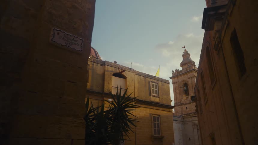 A slow dolly shot reveals an ancient church with a red dome and bell tower from a shadowy stone alley in Malta