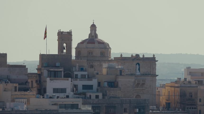 A historic Maltese cityscape featuring a grand cathedral with a dome and bell tower under a bright, hazy sky