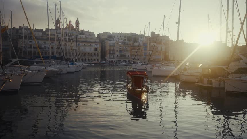 A traditional boat is rowed through a historic harbor in Malta, with sailboats and the rising sun creating a serene cityscape view