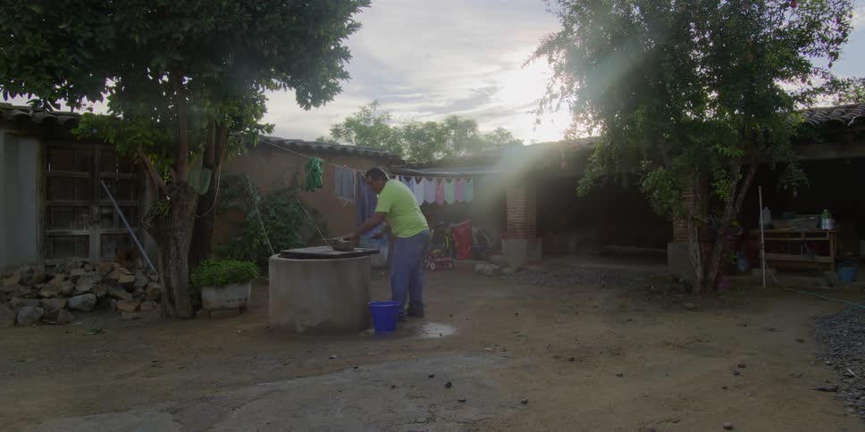 A man in a lime green shirt washes dishes over a well in the rustic courtyard of a home at sunset