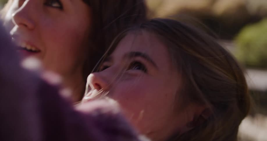 A young girl and her mother cuddle under a blanket, looking up at the sky with faces full of wonder and delight