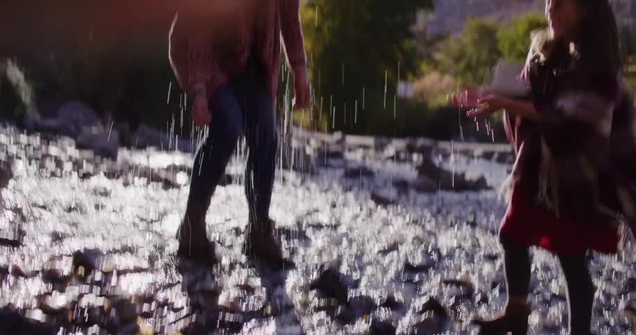 A young girl playfully splashes her laughing mother in a sunlit rocky creek in slow motion