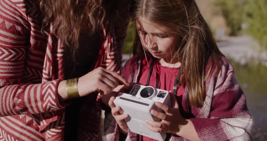 A mother shows her young daughter how to use a white instant camera while spending time together outdoors on a sunny day