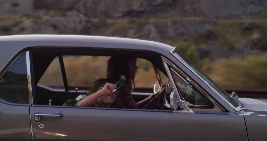 A smiling girl in sunglasses takes selfies and photos with an instant camera while on a road trip with her friend in a classic car