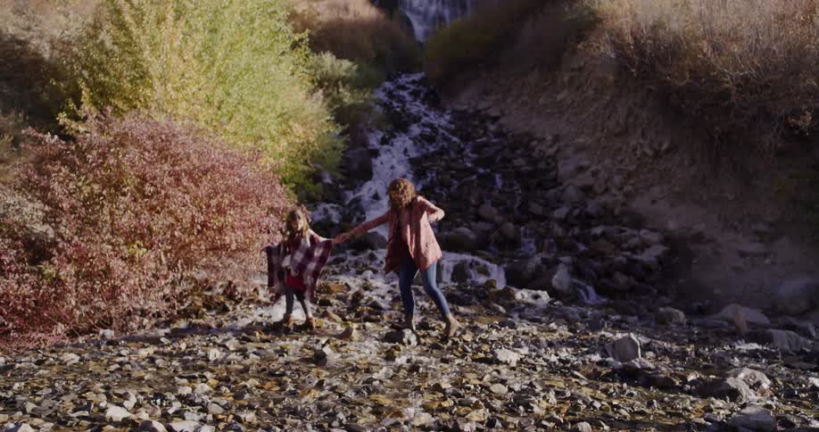 Slow motion tilt down from a large waterfall to two young women holding hands and playing on the rocky riverbed below