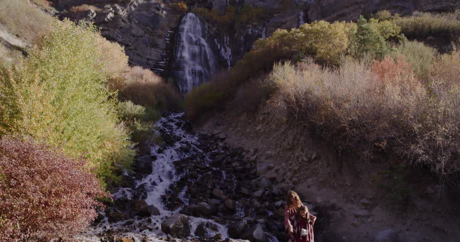 A mother and daughter share a warm moment by a rocky stream as the camera tilts up to reveal a majestic waterfall in a beautiful autumn landscape