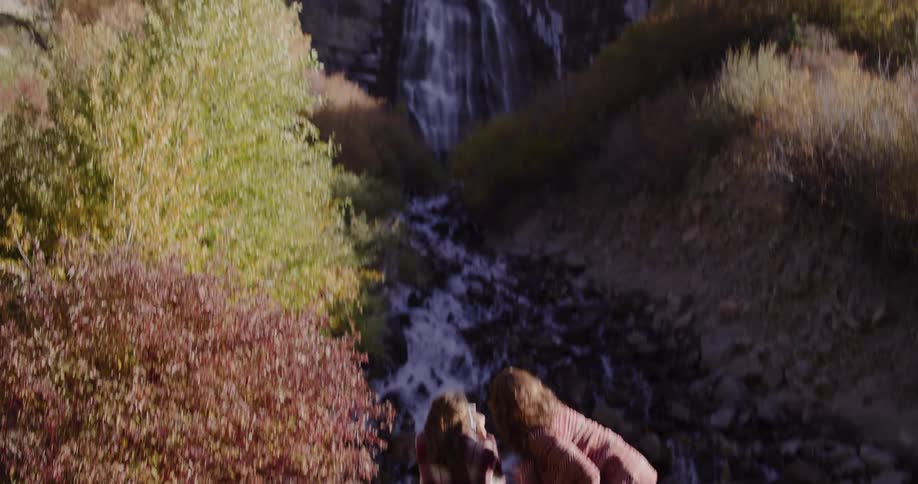 A mother and daughter stand at the base of a large waterfall in a mountain canyon, with the camera tilting up to reveal the full scenic view