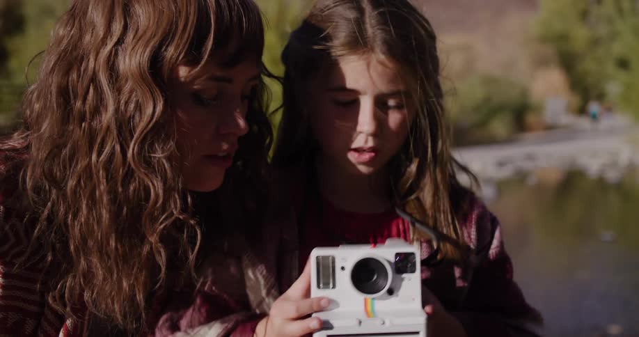 A mother teaches her young daughter how to use a vintage-style instant camera while wrapped together in a cozy blanket by a river