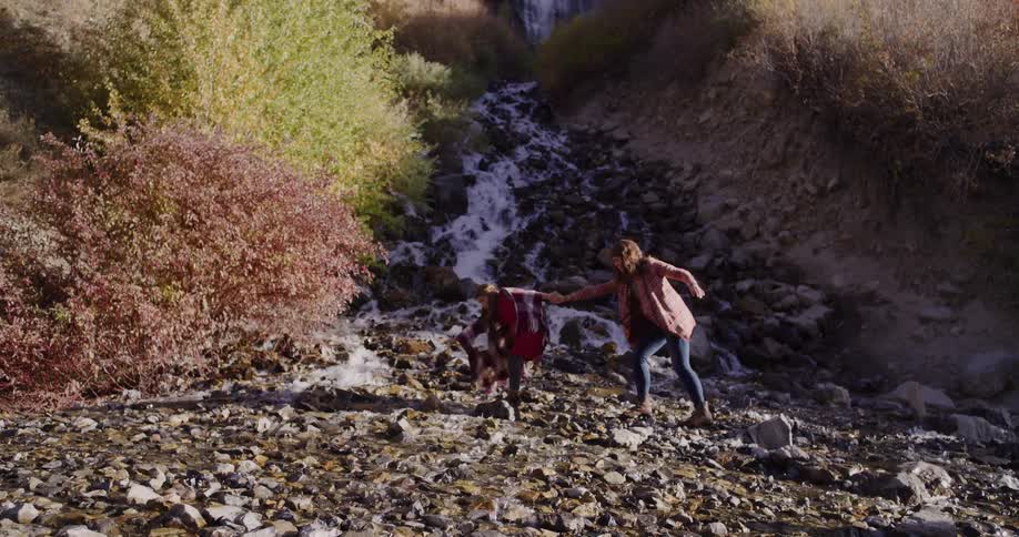 A mother holds her daughter's hand as they carefully cross a rocky stream below a scenic waterfall in the mountains