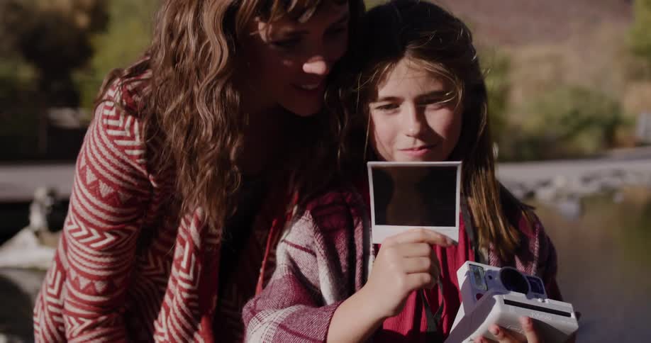 A smiling girl and her mother watch a polaroid picture develop while enjoying a sunny day together in nature