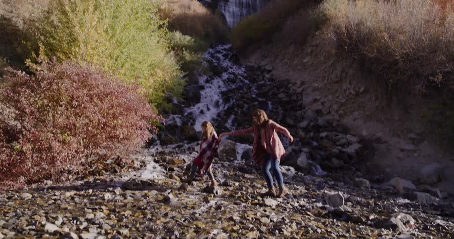 A wide shot of a beautiful mountain waterfall tilts down to reveal a mother and daughter holding hands while crossing a rocky stream below