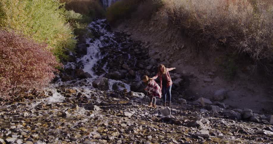 A mother and daughter carefully walk along the rocks of a mountain stream in slow motion on a beautiful autumn day