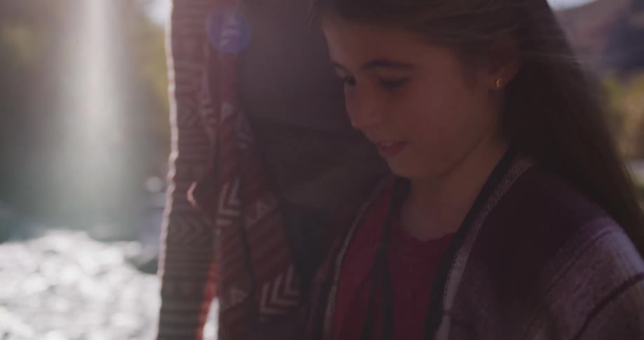 A happy young girl watches as her instant camera prints a photo in slow motion while standing by a sunny river