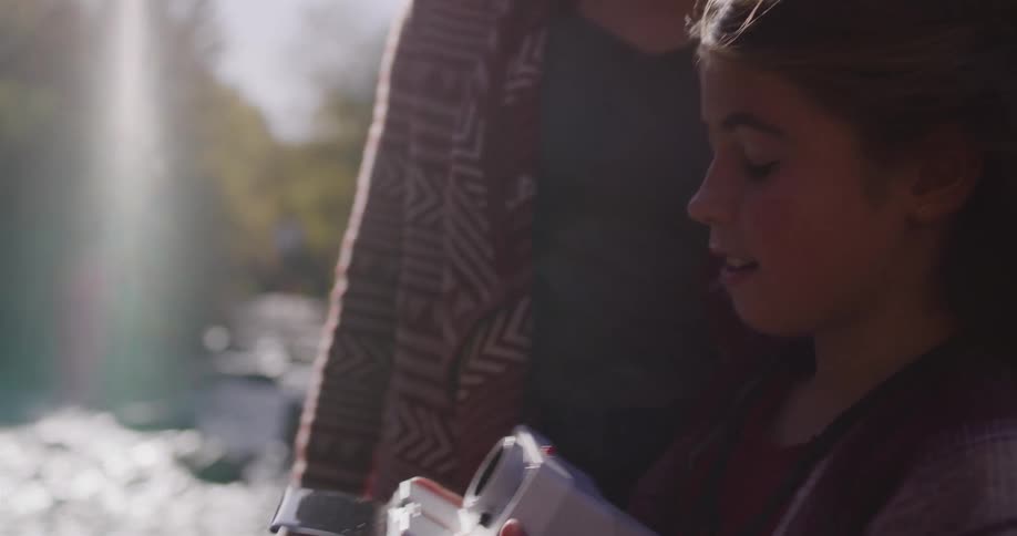 A smiling young girl takes a picture with an instant camera and watches it develop in slow motion by a sunlit river