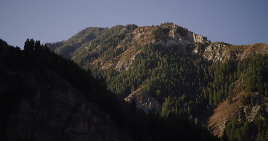 A slow zoom-out reveals a majestic, sunlit mountain peak towering over a shadowed, tree-covered valley under a clear blue sky