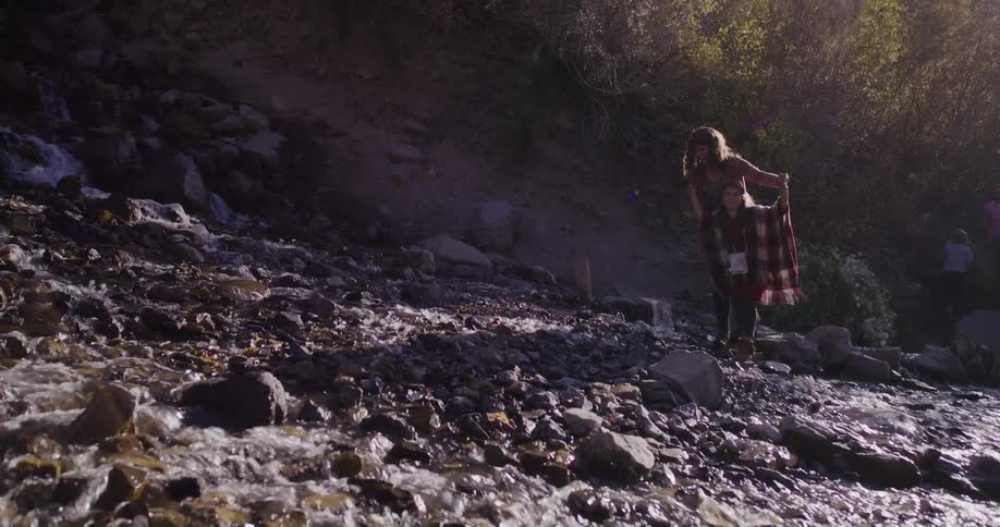 Mother gives her daughter a piggyback ride while blowing bubbles next to a mountain stream in slow motion