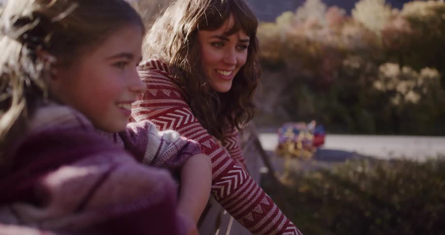 A mother and daughter share a loving moment together, smiling and talking on an outdoor railing on a sunny autumn day