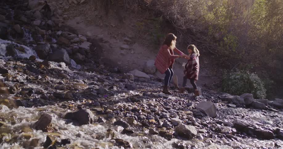 A mother carefully guides her young daughter across a rocky mountain stream, holding her hands for support in slow motion