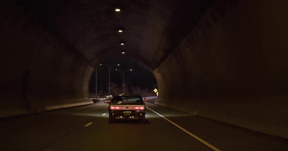 A classic car drives through a mountain canyon tunnel at dusk, with a passenger's arms stretched out in freedom