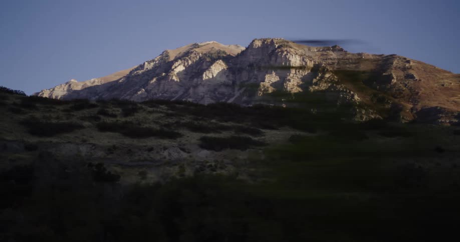A view of a rugged, sunlit mountain range with blurred trees passing in the foreground, shot from a moving vehicle