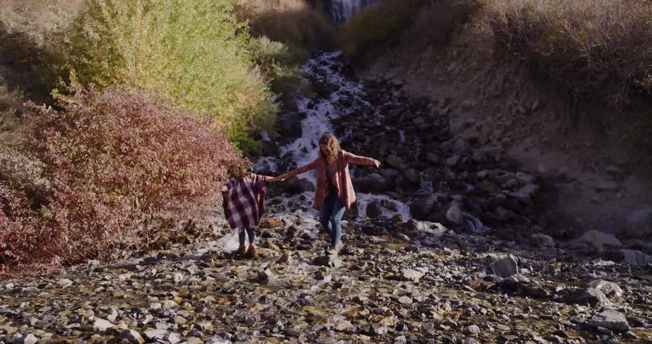 In slow motion, a woman holds a young boy's hand, guiding him as they carefully cross a rocky mountain stream in autumn