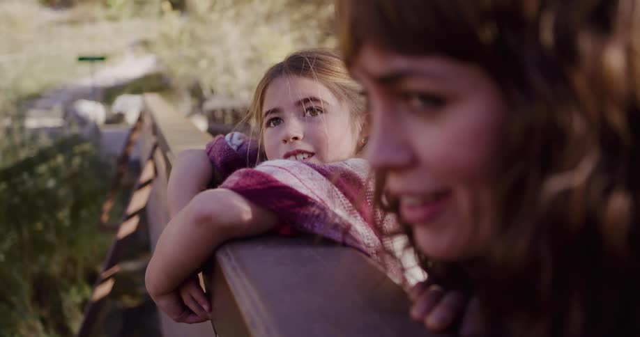 A daughter wearing a poncho talks with her mother while they both lean over a bridge railing in a sunny park