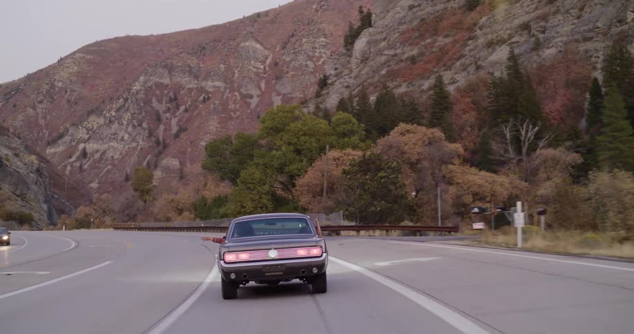 A person hangs their arms out the windows of a classic muscle car while driving through a scenic mountain canyon road at dusk