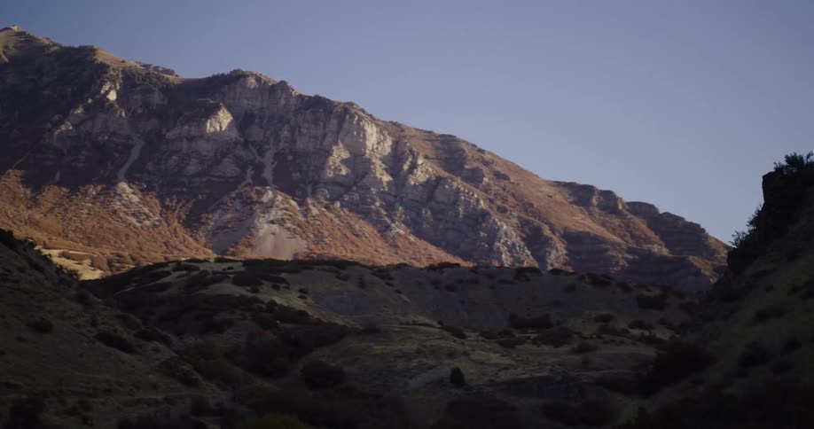 A slow pan reveals the rugged, sunlit peaks of a mountain range contrasting with the shadowy hills in the foreground