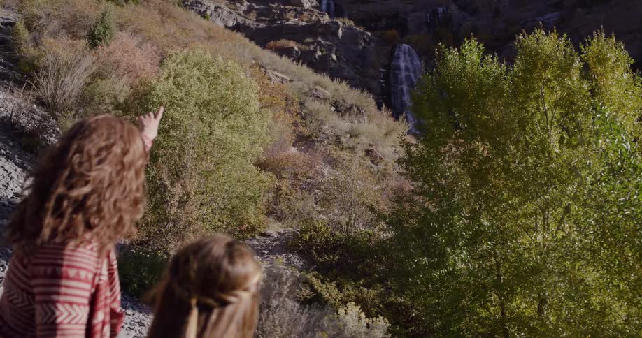 Over the shoulder shot of a mother and daughter pointing at a beautiful waterfall while on a hike in the mountains