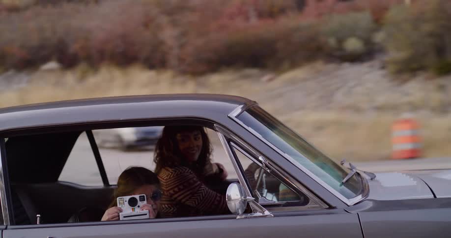 A young girl takes a picture with an instant camera from the passenger window of a classic car while her friend drives down a highway