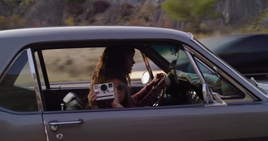 A young girl with an instant camera enjoys a road trip with her mother in a classic car through the countryside