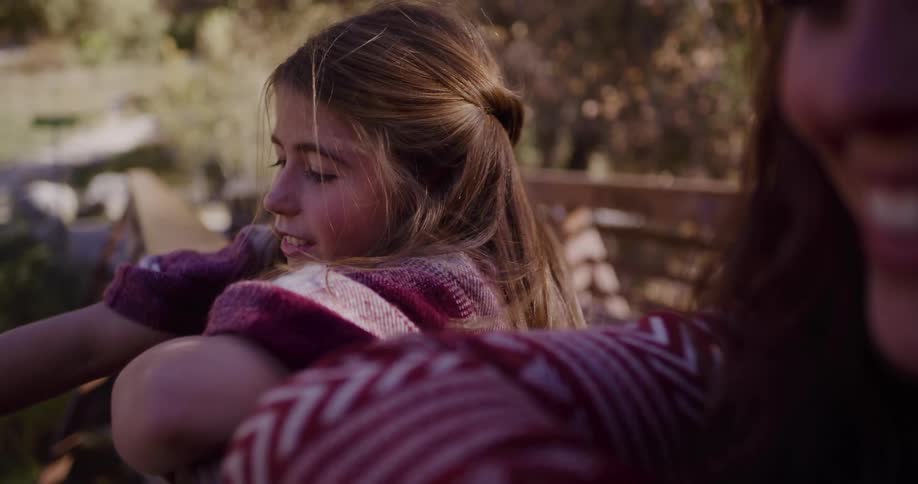 A young girl in profile leans on a railing next to her mother, her hair blowing in the wind during a quiet outdoor moment