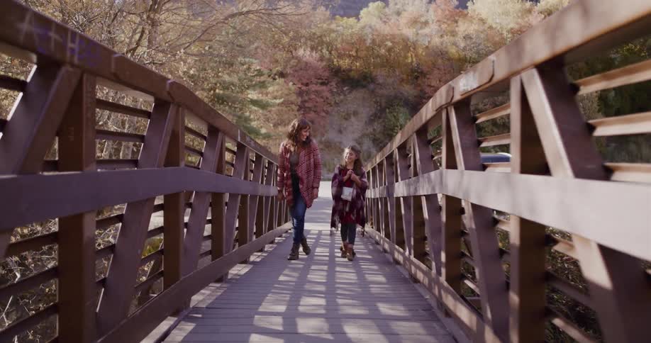 A mother and her daughter smile and talk while walking across a wooden bridge on a beautiful autumn day