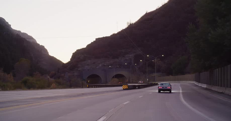 A classic car drives along a scenic highway through a mountain canyon, approaching a pair of tunnels at dusk