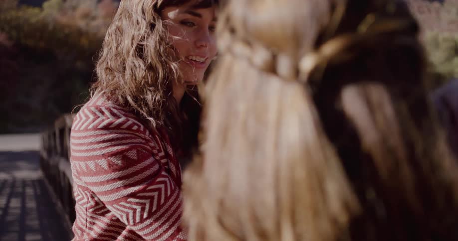 A close-up slow-motion shot captures two young women having an intimate conversation on a windy bridge in a sunlit natural setting