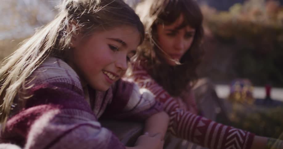 A young girl smiles warmly as a woman looks on with surprise and wonder while they lean over a railing outdoors in the sun