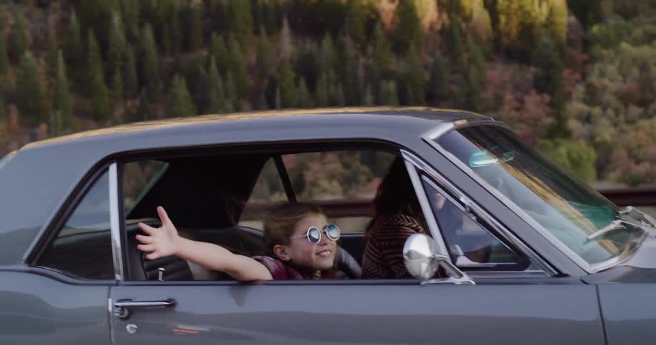Carefree young girl with retro sunglasses smiles and waves her arm out the window of a classic car on a scenic mountain road trip with her mother