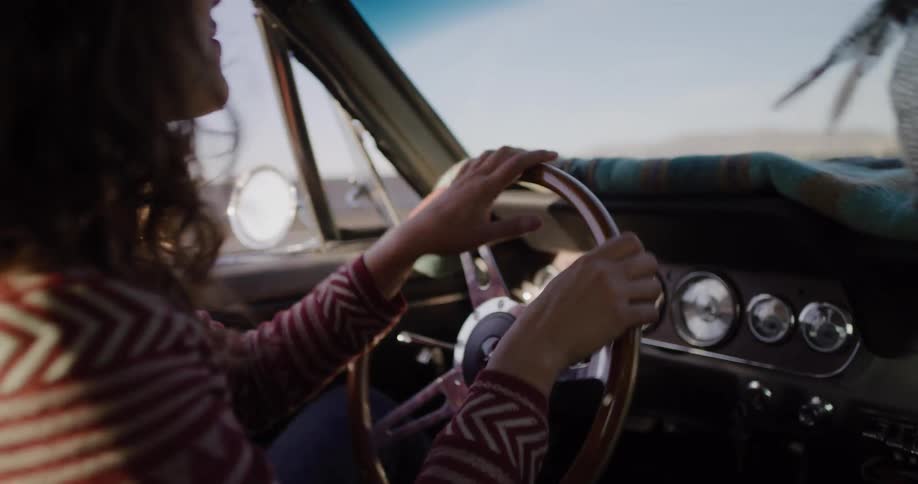 A woman in a southwestern-style sweater enjoys a sunny drive, tapping her hands on the steering wheel of her vintage car