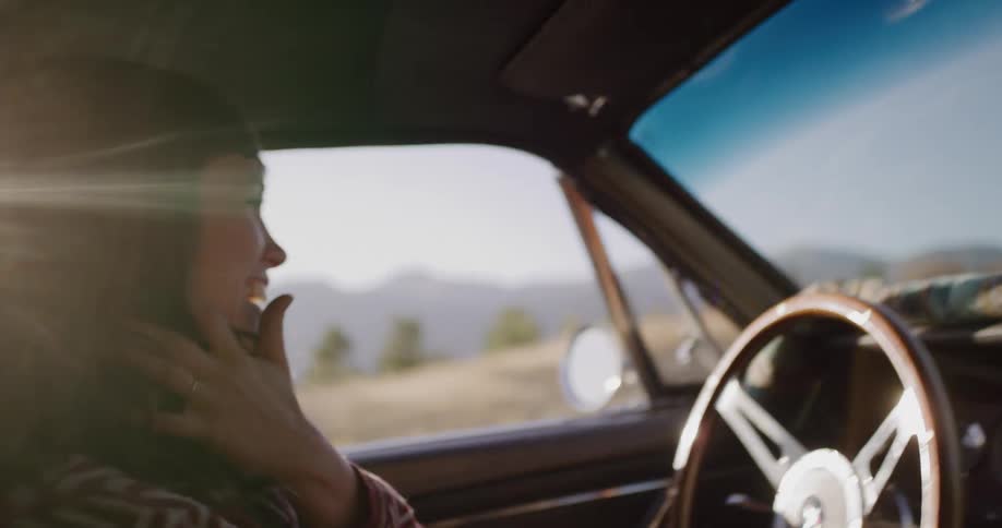 A happy woman smiles and laughs while driving a classic car on a sunny road trip, her hair blowing in the wind in slow motion
