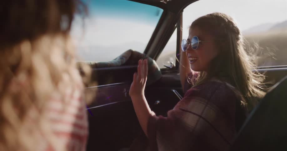 A carefree young woman in sunglasses smiles and dances to the music in the passenger seat of a vintage car during a sunny road trip