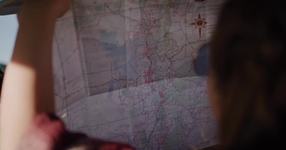 From the passenger seat, a woman points to a location on a physical map while the driver enjoys a soda in their classic car on a sun-drenched road trip