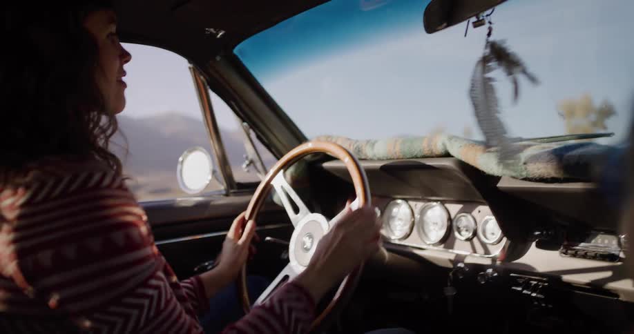 A carefree woman dances and sings along to the music while driving her vintage muscle car down a sunny road