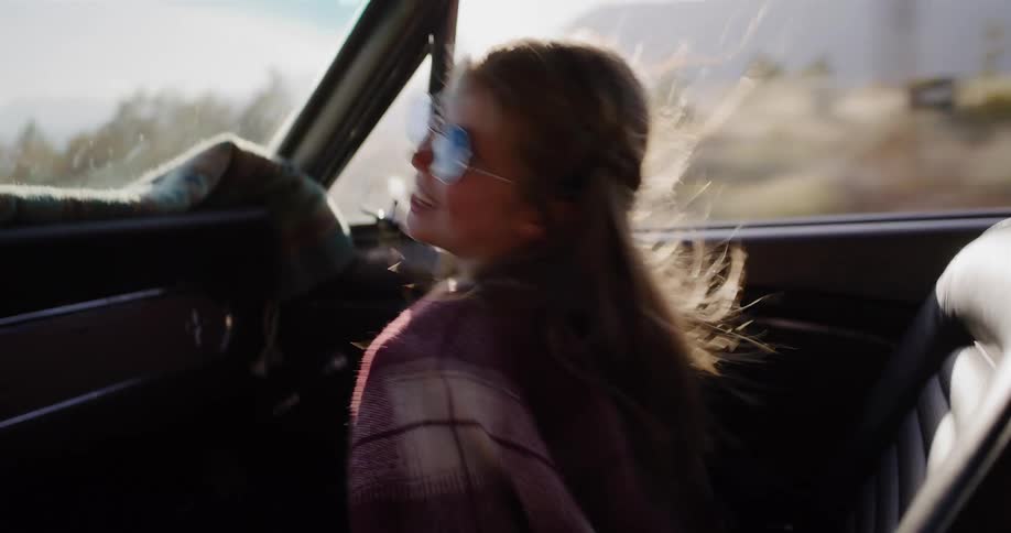 A happy young girl in retro sunglasses enjoys a scenic road trip in a classic convertible car at sunset