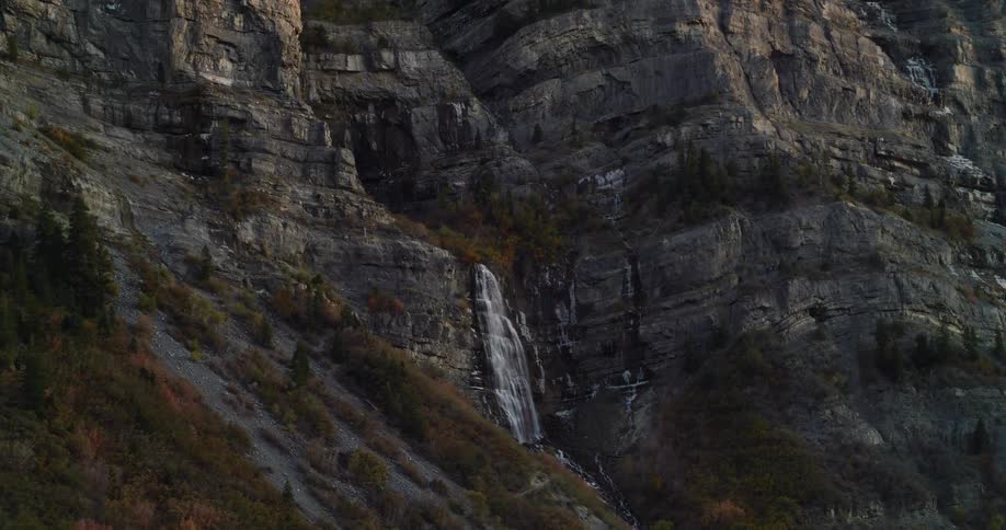 A majestic waterfall flows down the rugged face of a mountain surrounded by autumn foliage at sunset